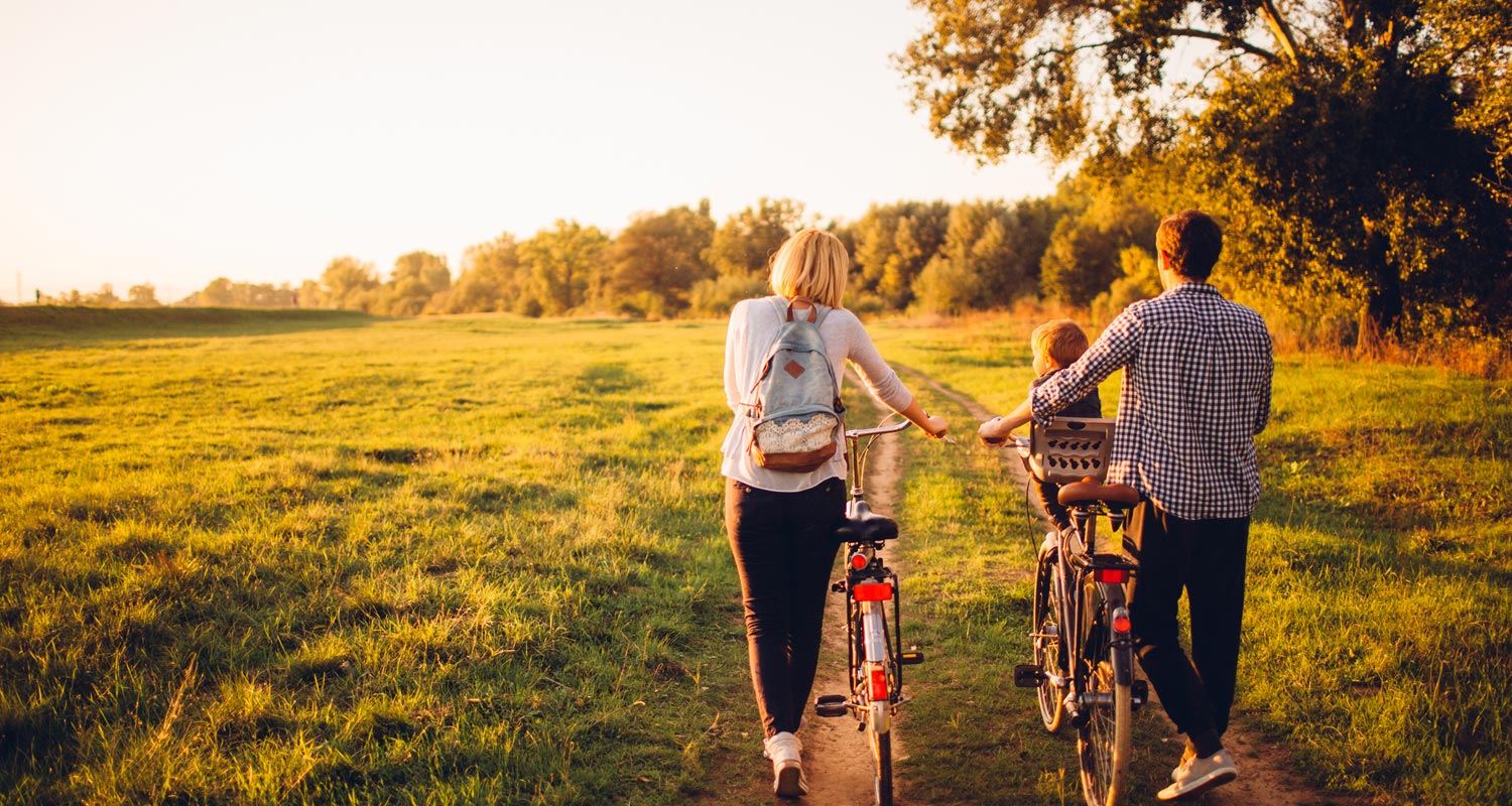 Two people pushing bikes through a field with a child sat in a seat on one of the bikes