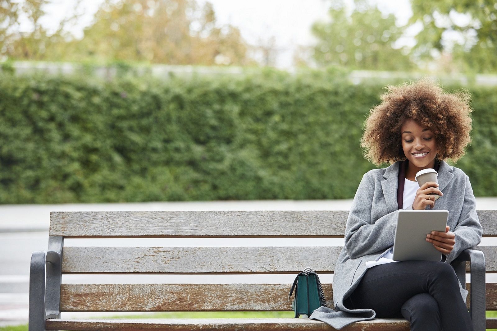 Photo of a woman sat on a park bench with a coffee and a tablet