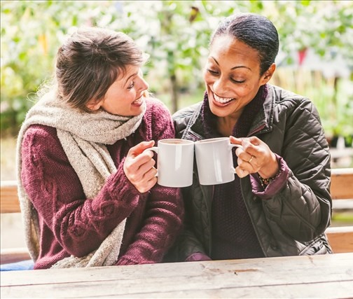 2 women drinking mugs of tea together at a table in a public park